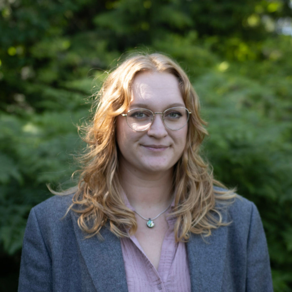 Portrait of a smiling woman with glasses wearing a gray blazer outdoors, green foliage in the background