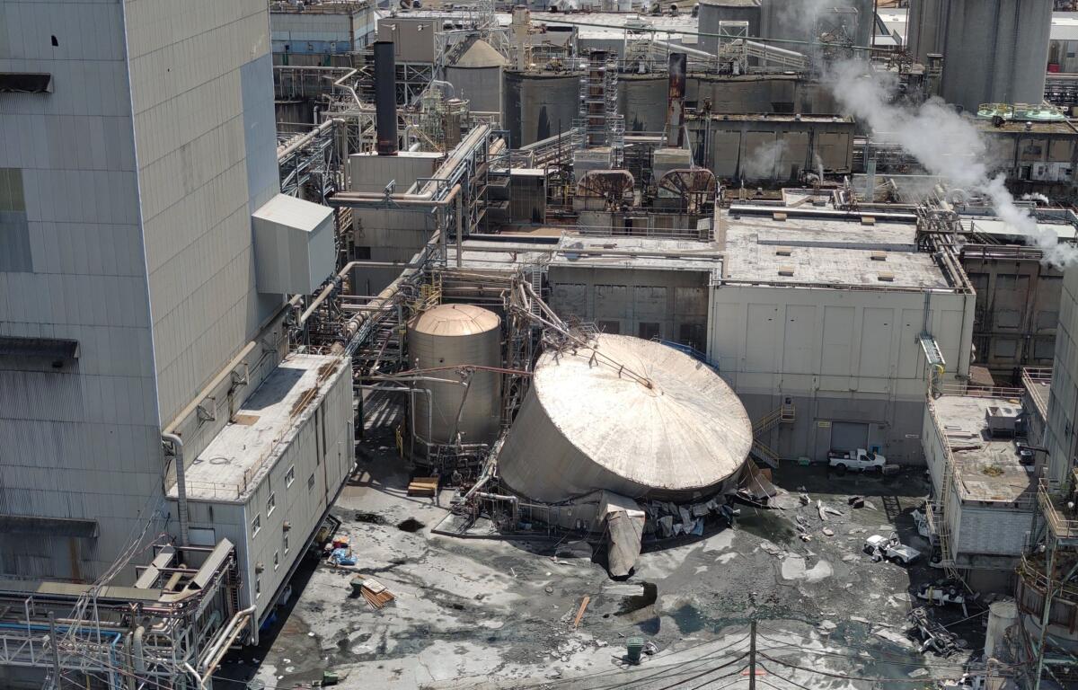 Aerial view of a large industrial plant with connected pipes, tanks, and smokestacks emitting steam. In the background are more structures and vehicles in a yard.