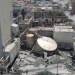 Aerial view of a large industrial plant with connected pipes, tanks, and smokestacks emitting steam. In the background are more structures and vehicles in a yard.