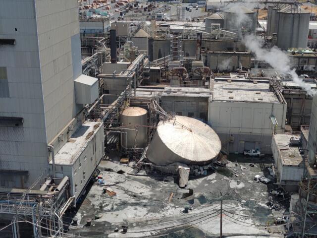 Aerial view of a large industrial plant with connected pipes, tanks, and smokestacks emitting steam. In the background are more structures and vehicles in a yard.