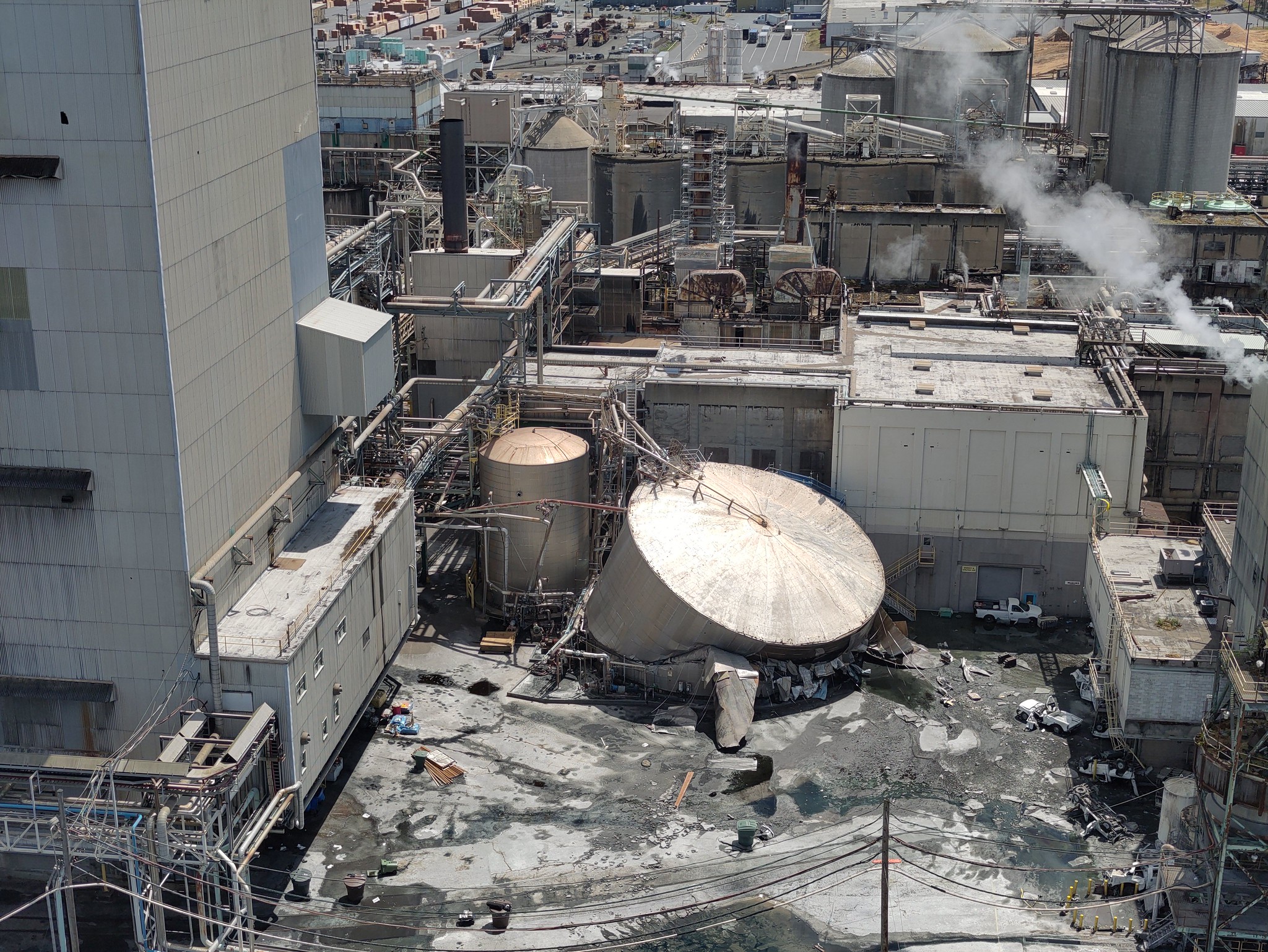 Aerial view of a large industrial plant with connected pipes, tanks, and smokestacks emitting steam. In the background are more structures and vehicles in a yard.