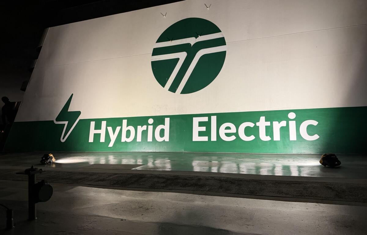 Large banner reading 'Hybrid Electric' with a green circular logo, stage floor and spotlights casting shadows.