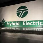Large banner reading 'Hybrid Electric' with a green circular logo, stage floor and spotlights casting shadows.