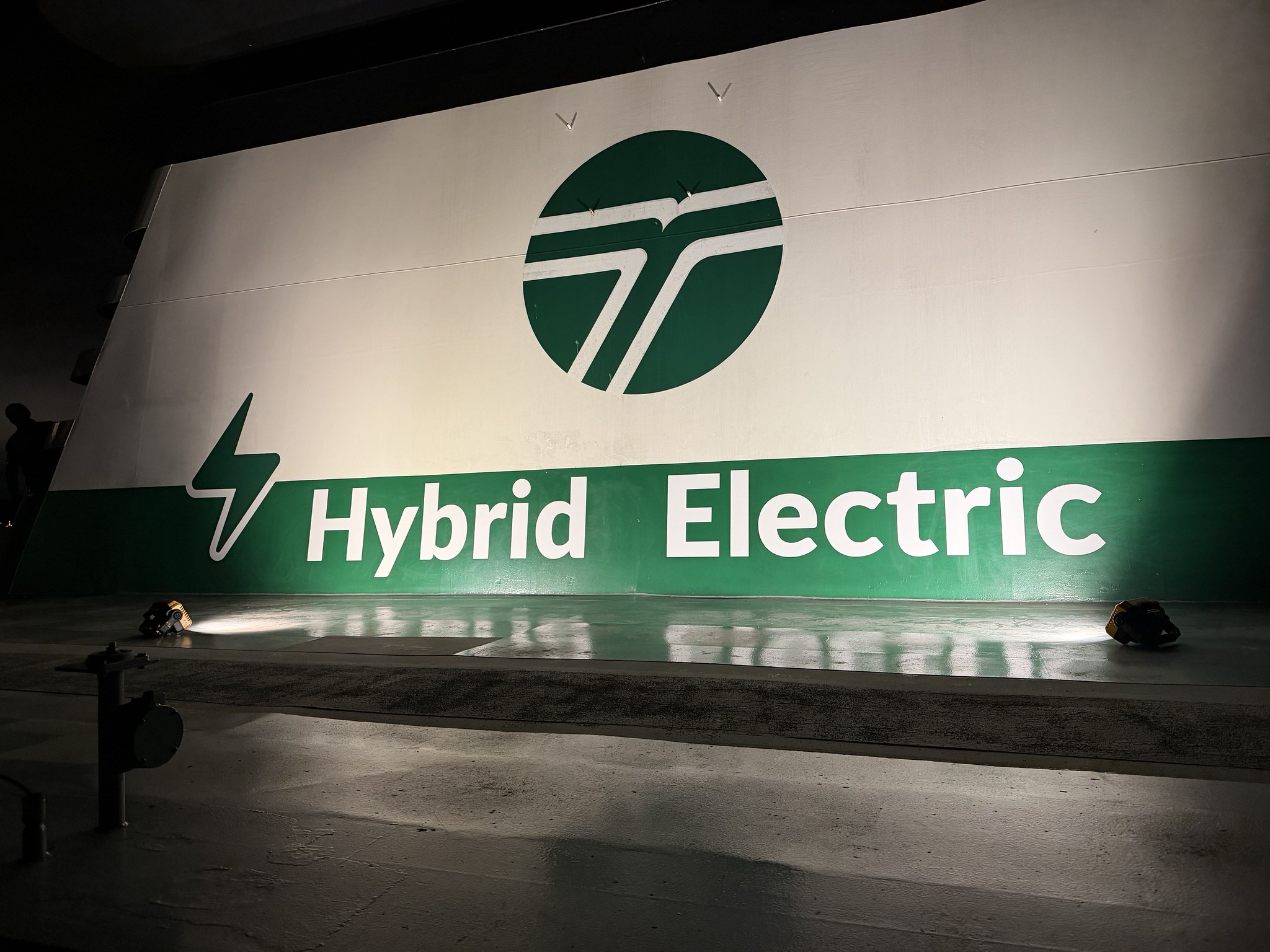 Large banner reading 'Hybrid Electric' with a green circular logo, stage floor and spotlights casting shadows.