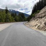 Curving mountain road flanked by a rocky cliff on the right and dense evergreen forest on the left, with snow-capped mountains in the distance.