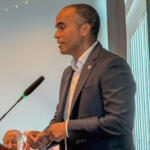 Man in a dark suit giving a speech at a podium with two microphones in front of him.