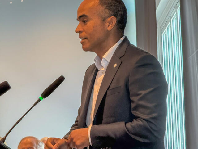 Man in a dark suit giving a speech at a podium with two microphones in front of him.