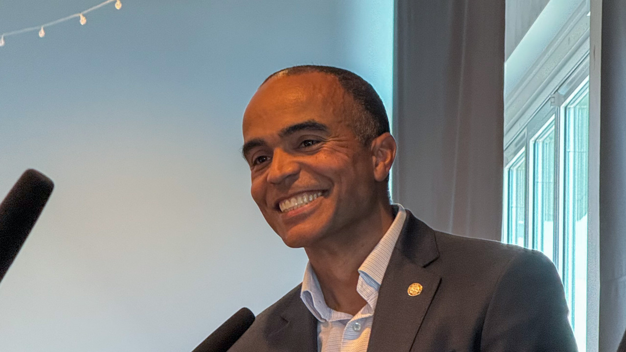 Smiling man in a dark suit speaks at a podium with two microphones in front of him.