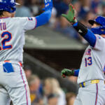 Two Mets players share a high-five on the field, Soto wearing #22 and a teammate #13 nearby against a blurred crowd backdrop.