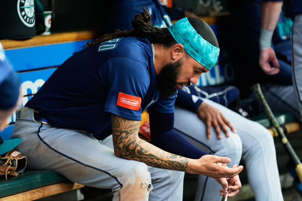 Baseball player sitting on a dugout bench, with a teal headband and Nintendo patch on his navy jersey, examining his hands momentarily.
