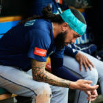Baseball player wearing a teal headband and blue jersey sits on a dugout bench, examining a small object in his hands, with tattoos on his arm.