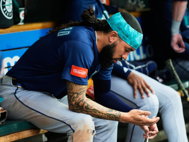 Baseball player wearing a teal headband and blue jersey sits on a dugout bench, examining a small object in his hands, with tattoos on his arm.