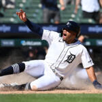 Detroit Tigers player sliding into a base, kicking up dirt with his arm raised.