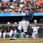 Baseball batter in a dark blue uniform swings at a pitch at home plate with a blurred crowd in the stands behind him.