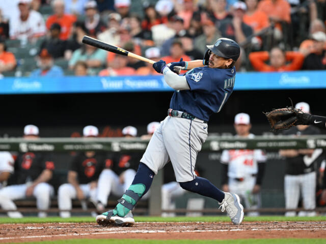 Baseball batter in a dark blue uniform swings at a pitch at home plate with a blurred crowd in the stands behind him.