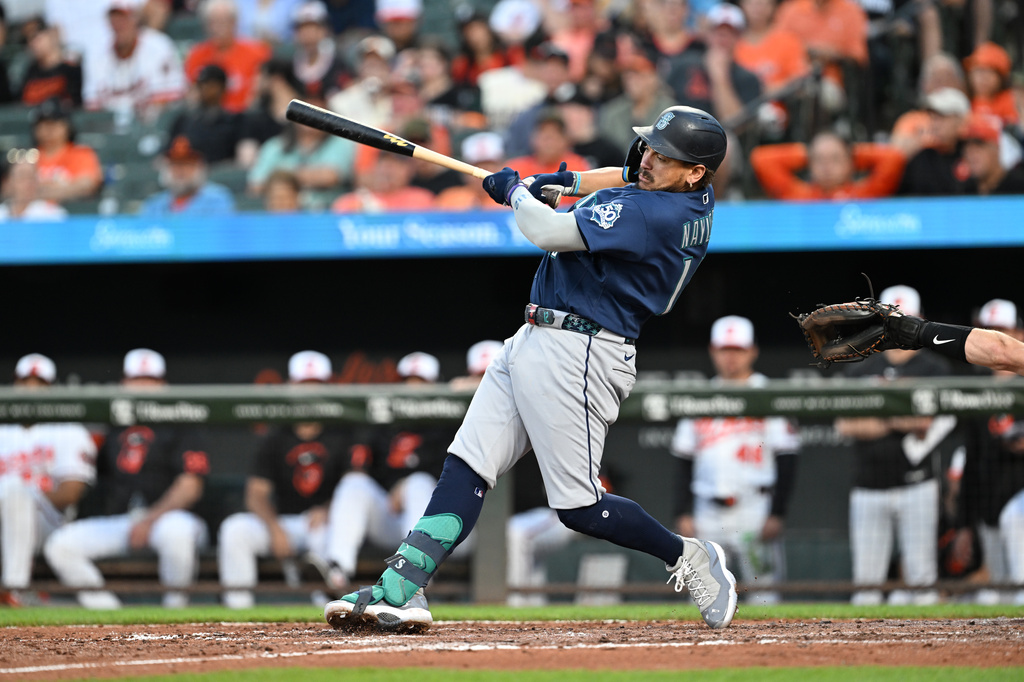 Baseball batter in a dark blue uniform swings at a pitch at home plate with a blurred crowd in the stands behind him.
