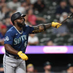 Seattle Mariners batter in blue uniform swings at a pitch during a game, wearing yellow batting gloves.