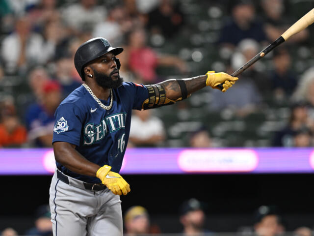 Seattle Mariners batter in blue uniform swings at a pitch during a game, wearing yellow batting gloves.