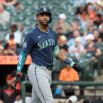 Seattle Mariners batter in navy uniform and helmet walking on the field during a game, crowd in the stands behind him.
