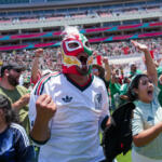 Crowd of soccer fans at a stadium, center figure wearing a colorful lucha libre mask and white Mexico jersey, arms raised in cheer.