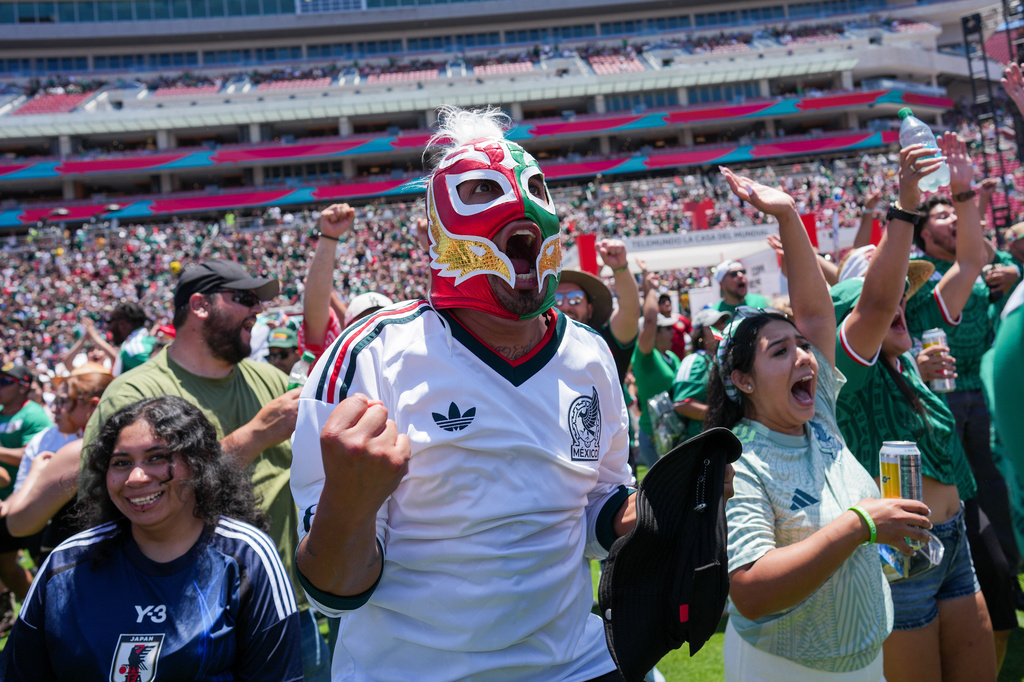 Crowd of soccer fans at a stadium, center figure wearing a colorful lucha libre mask and white Mexico jersey, arms raised in cheer.