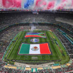 Aerial view of a packed stadium with two large flags unfurled on the field and colorful smoke rising above the stands.