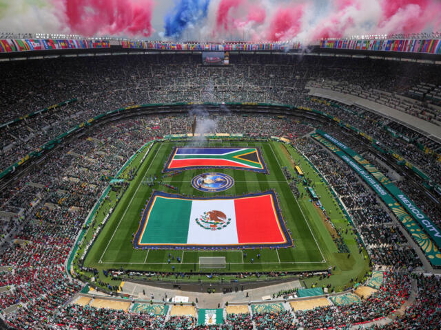 Aerial view of a packed stadium with two large flags unfurled on the field and colorful smoke rising above the stands.