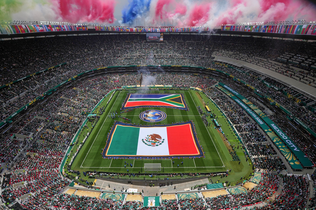 Aerial view of a packed stadium with two large flags unfurled on the field and colorful smoke rising above the stands.