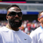 Two men at a sports stadium wearing white shirts and sunglasses, one with a gold chain, blurred crowd in the background.