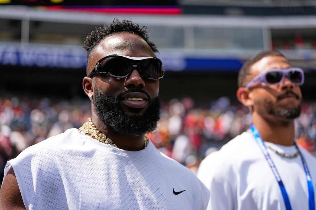 Two men at a sports stadium wearing white shirts and sunglasses, one with a gold chain, blurred crowd in the background.