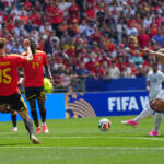 White-team player kicks the ball toward goal as red-clad defenders approach in a FIFA World Cup match, stadium packed with fans.