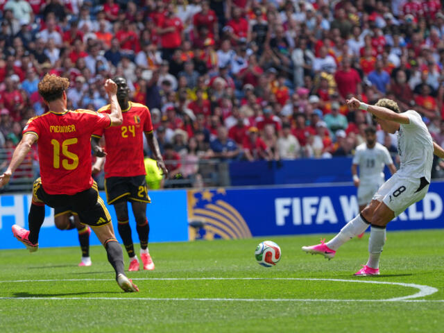 White-team player kicks the ball toward goal as red-clad defenders approach in a FIFA World Cup match, stadium packed with fans.