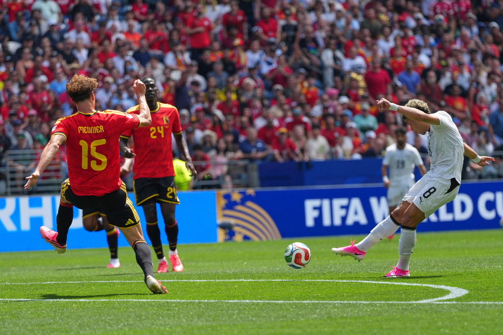White-team player kicks the ball toward goal as red-clad defenders approach in a FIFA World Cup match, stadium packed with fans.