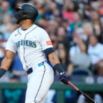 Seattle Mariners batter in white uniform and helmet swings a bat during a game, crowd in the stands behind him.