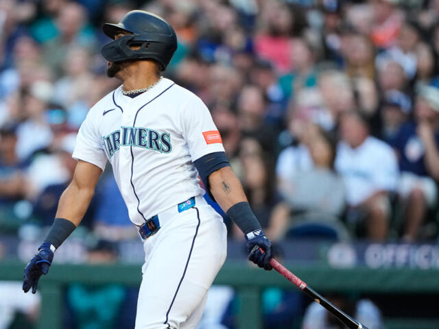 Seattle Mariners batter in white uniform and helmet swings a bat during a game, crowd in the stands behind him.