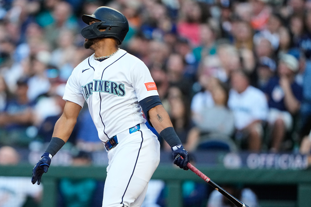 Seattle Mariners batter in white uniform and helmet swings a bat during a game, crowd in the stands behind him.