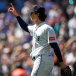 Mariners pitcher in white uniform and cap raises his right arm, index finger pointing, with a blurred crowd in the background.