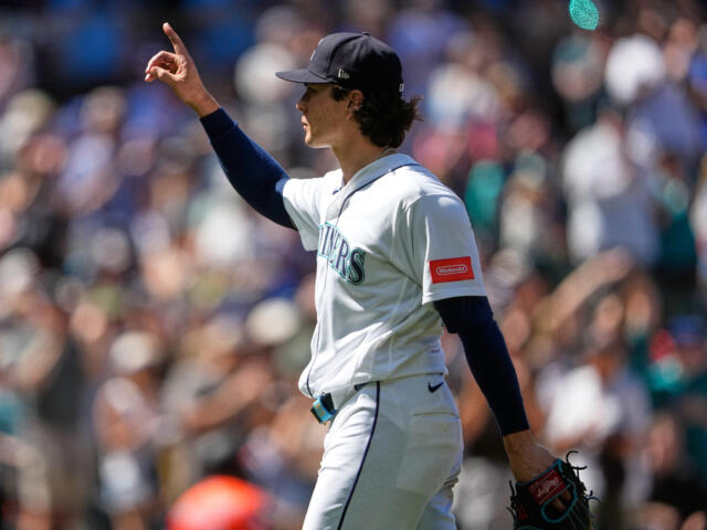 Mariners pitcher in white uniform and cap raises his right arm, index finger pointing, with a blurred crowd in the background.