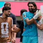 Post‑match celebration as a goalkeeper in blue hugs a teammate, with another player wearing a brown FIFA World Cup 2026 bib looking on in the foreground.