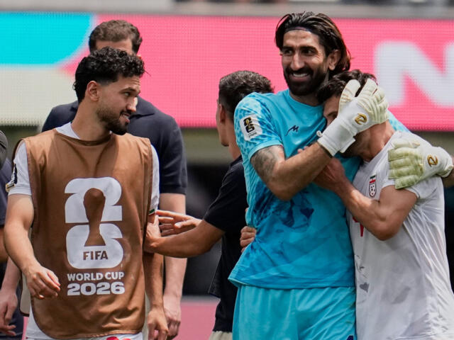 Post‑match celebration as a goalkeeper in blue hugs a teammate, with another player wearing a brown FIFA World Cup 2026 bib looking on in the foreground.