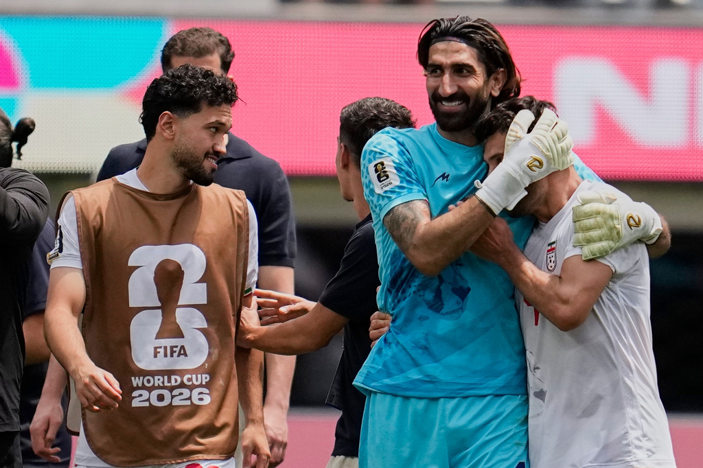 Post‑match celebration as a goalkeeper in blue hugs a teammate, with another player wearing a brown FIFA World Cup 2026 bib looking on in the foreground.