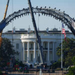 Large steel arch structure under construction in front of the White House, with a US flag nearby and workers on site.