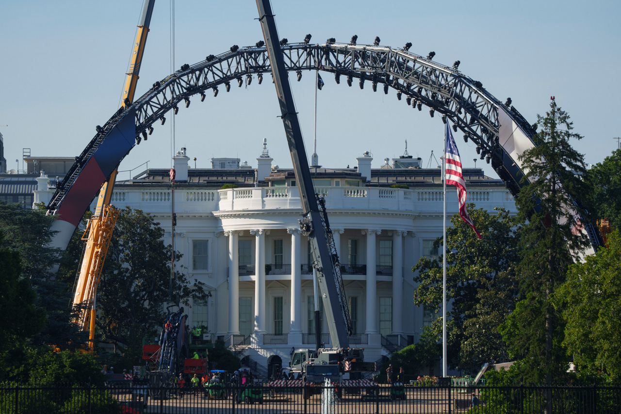 Large steel arch structure under construction in front of the White House, with a US flag nearby and workers on site.
