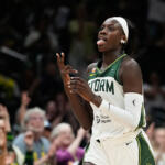 Female basketball player in a white and green Storm jersey on the court, sticking out her tongue as she moves.