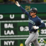 Seattle Mariners player in a navy jersey runs with his right arm raised in celebration on a baseball field, green outfield wall behind him.