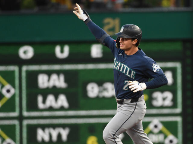 Seattle Mariners player in a navy jersey runs with his right arm raised in celebration on a baseball field, green outfield wall behind him.