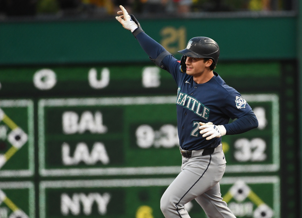 Seattle Mariners player in a navy jersey runs with his right arm raised in celebration on a baseball field, green outfield wall behind him.