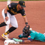 Pirates infielder in black and yellow stretches to tag a teal-uniformed runner sliding into a base; ball in midair near the glove at the base.