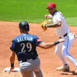 Baseball action: fielder in white throws as Raleigh 29 in blue watches near the base path on the dirt infield.