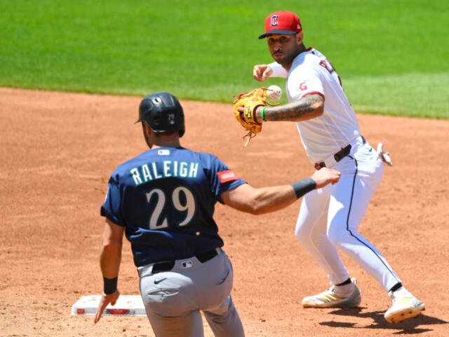 Baseball action: fielder in white throws as Raleigh 29 in blue watches near the base path on the dirt infield.